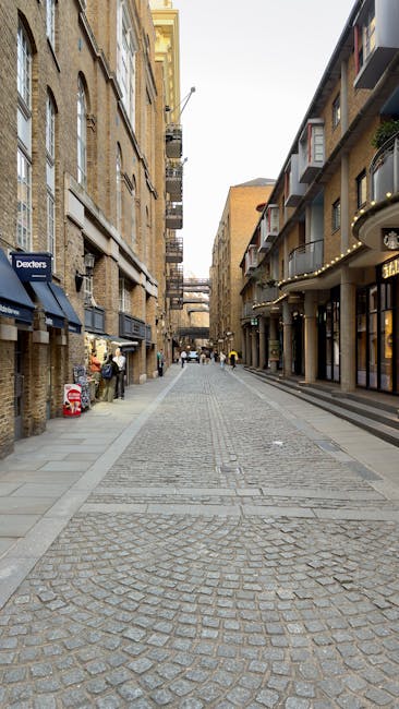 A cobbled street scene in Hampstead with multi-storey buildings lining both sides, featuring a mix of brick and modern facades. On the left side, there are shops with dark blue awnings, one displaying a sign for 'Dexters,' and adjacent to it, a person standing next to a stack of large, flat cardboard boxes and packing materials. On the right side, there are residential and commercial buildings with balconies, some featuring modern glass and metal façades. In the foreground, there are loading areas with open doorways where items such as plastic-wrapped furniture or large household boxes might be temporarily stored during a home or office relocation process. The street is under daylight with natural light illuminating the scene, and the background shows several pedestrians walking, with a visible construction crane indicating ongoing development. Hampstead Removals collaborates with local businesses and residents to facilitate furniture transport and packing during relocations, supporting smooth moving logistics within this urban area.