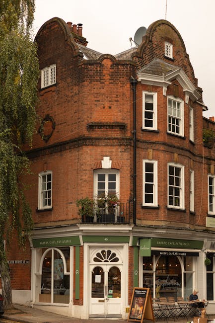 Photograph of a three-storey red brick building with a ground-floor shopfront and residential windows above, located on a city street. The shop has green signage and a white door with a decorative semi-circular window, adjacent glass windows with arched tops, and a small sandwich board outside. The upper floors feature white-framed sash windows, some with small balconies and flowerpots, set within curved brickwork at the building's corner and roofline. A person sits on a bench on the pavement nearby, while a large tree partially obscures the left side of the building. Visible in the background are satellite dishes and chimney stacks on the roof. This scene reflects typical urban property with mixed-use ground-floor commercial space and residential accommodation above, illustrating building features relevant to house and commercial removals, packing, and furniture transport processes. Hampstead Removals regularly facilitates such local relocations, ensuring careful handling of furniture and belongings during loading and transport activities.