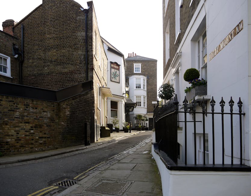 A narrow residential street in Hampstead with a slight uphill incline, featuring a mix of traditional brick buildings and modern white facades. On the right, a white building with large windows and decorative black wrought iron railings, alongside potted plants with green foliage and purple flowers. An area of the pavement near the building displays several cardboard boxes, packing materials, and a small wooden stool, indicating a home relocation process. On the left, a brick building with a dark roof and a wall at street level, partially obscuring the view down the winding street. In the background, additional narrow streets, more residential buildings with bay windows, and chimneys are visible under natural daylight, suggesting daylight hours. The scene captures a typical London neighbourhood environment suitable for houses removals and furniture transport, with the Hampstead Removals service potentially involved in managing the logistics of packing and moving objects through tight streets and on stairs.