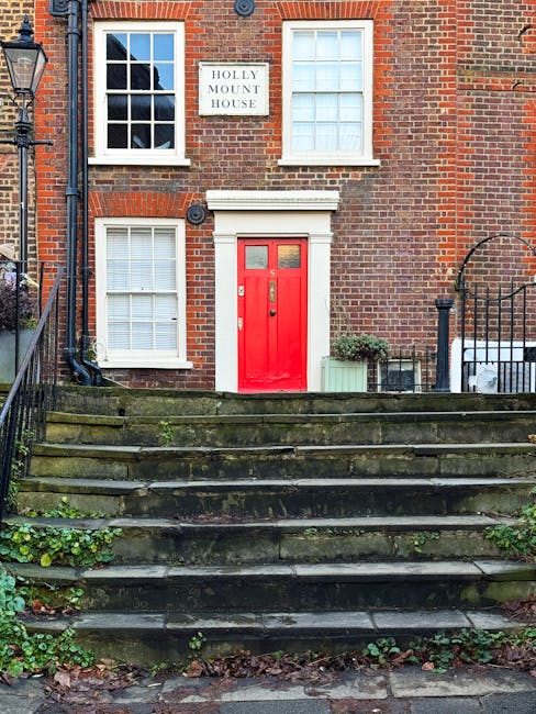 A brick residential building with a white sign reading 'Holly Mount House' mounted on the upper part of the facade. The building features white-framed sash windows on both the ground and first floors, with white window sills. The entrance has a vibrant red door set into a white doorframe, accessible via a short flight of moss-covered stone steps bordered by black wrought iron railings. To the right of the door, there is a small green potted plant on a ledge, with a green planter box nearby. The scene is lit by natural daylight, highlighting the textures of the brickwork, the moss on the stairs, and the surrounding greenery, which includes some low-lying plants and ivy at the bottom of the steps. The setting suggests a typical historic London house, and the image may be used in contexts related to house removals, packing, or home relocation services, such as Hampstead Removals, particularly for narrow streets and stairs.