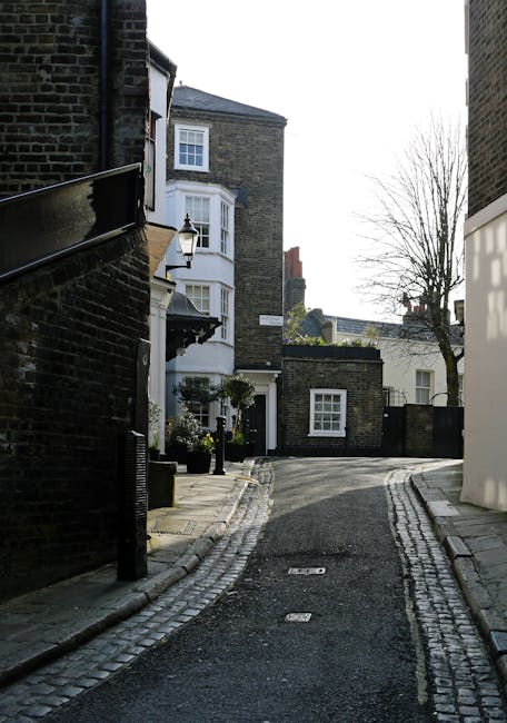 A narrow residential street in Hampstead with a slight uphill incline, featuring a mix of traditional brick buildings and modern white facades. On the right, a white building with large windows and decorative black wrought iron railings, alongside potted plants with green foliage and purple flowers. An area of the pavement near the building displays several cardboard boxes, packing materials, and a small wooden stool, indicating a home relocation process. On the left, a brick building with a dark roof and a wall at street level, partially obscuring the view down the winding street. In the background, additional narrow streets, more residential buildings with bay windows, and chimneys are visible under natural daylight, suggesting daylight hours. The scene captures a typical London neighbourhood environment suitable for houses removals and furniture transport, with the Hampstead Removals service potentially involved in managing the logistics of packing and moving objects through tight streets and on stairs.
