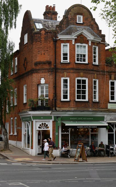 The image depicts a three-storey red brick period building located on Heath Street, Hampstead, with a ground-floor bakery, café, and pâtisserie featuring a green awning and large glass windows. The upper floors have white-framed sash windows, some with small balconies, and a curved gabled roof with skylights. In front of the shop, a sidewalk scene includes three people walking past, two seated customers at outdoor tables, and a blackboard sign on the pavement. The scene is set during the daytime with natural light, and trees partially shade the building on the left. The image is relevant to house removals and relocation services provided by Hampstead Removals, illustrating the typical street environment involved in local home relocations, furniture transport, and packing processes for properties in historic Hampstead.
