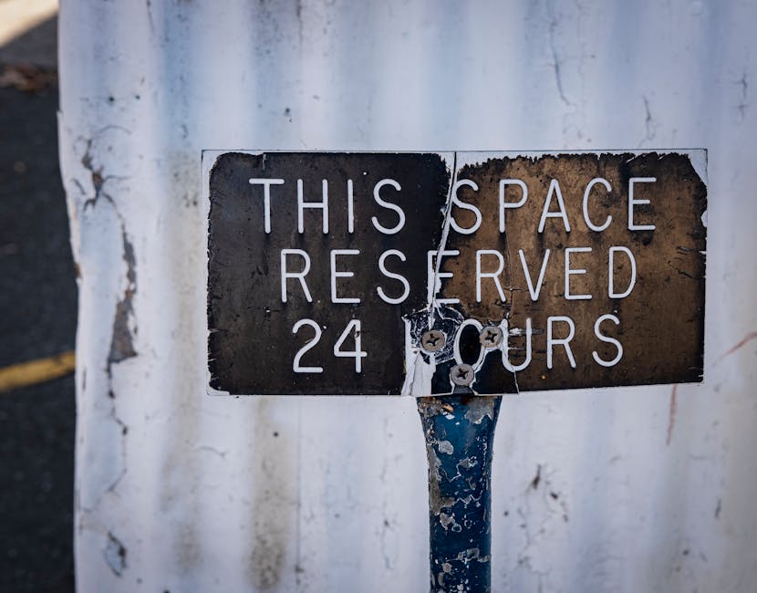 A weathered metal sign mounted on a post in front of a whitewashed wall, indicating a parking restriction with the message 'This space reserved 24 hours'. The sign shows signs of rust, peeling paint, and corrosion, with visible screws securing it to the post. The surrounding environment suggests an urban setting, relevant to house removals or relocation services such as those offered by Hampstead Removals. The sign's condition reflects typical street-side signage that may impact parking arrangements during a home relocation or furniture transport process, emphasizing the importance of understanding parking permits and access when planning NW3 flat removals. The overall scene captures the context of logistical considerations involved in loading and unloading within restricted parking zones, supporting careful planning for house moving services.