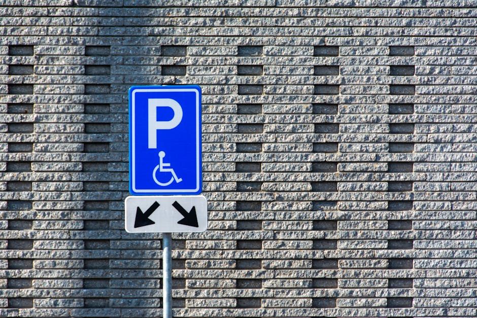 A blue parking sign with a white 'P' and a wheelchair symbol is mounted on a metal pole in front of a textured grey brick wall. Below the parking symbol, there are two black arrows pointing diagonally downward to the left and right, indicating accessible parking directions. The scene is well-lit with natural daylight, and the sign is positioned in an outdoor setting, likely near an entrance or parking area for a residential or commercial property. This image relates to access and parking regulations, which are important considerations during a home relocation or furniture transport, as highlighted in Hampstead Removals' NW3 flat removals advice for parking and permit access, ensuring efficient and compliant moving logistics.
