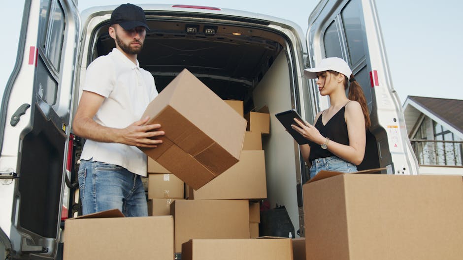 Inside the open back of a white delivery van, two individuals are engaged in the home relocation process. The man, wearing a beige cap, white polo shirt, and jeans, is holding a medium-sized cardboard box, preparing for furniture transport or packing and moving tasks. The woman, dressed in a white cap, black sleeveless top, and jeans, is standing nearby and using a tablet device, likely coordinating or managing the logistics of the move. The van's interior is filled with several cardboard boxes, some stacked, and a black plastic bag, all prepared for loading or unloading as part of a professional removals service by Hampstead Removals. The scene is set outdoors in a residential area, with a glimpse of a house with a pitched roof and a bay window in the background, suggesting work occurring during daylight hours. This image clearly depicts the process of packing, loading, and moving furniture and household items during a home relocation service.