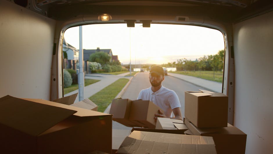 A man wearing a white shirt and sunglasses is standing outside a white moving van, holding a cardboard box during a home relocation process. The van's open rear doors reveal several packed cardboard boxes, some reinforced with packing tape and others with flaps open, positioned on the van's floor. The inside of the van is well-lit by natural sunlight, with visible mounting points and interior panels. Outside, a suburban street scene includes a sidewalk, a lamppost, and houses with front gardens, illuminated by the warm glow of the setting sun. The scene captures the logistics of furniture transport and packing during a house move, illustrating the loading process that Hampstead Removals handles as part of their specialist removals service, focusing on efficient and careful handling of items.