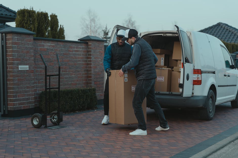 Inside the open back of a white delivery van, two individuals are engaged in the home relocation process. The man, wearing a beige cap, white polo shirt, and jeans, is holding a medium-sized cardboard box, preparing for furniture transport or packing and moving tasks. The woman, dressed in a white cap, black sleeveless top, and jeans, is standing nearby and using a tablet device, likely coordinating or managing the logistics of the move. The van's interior is filled with several cardboard boxes, some stacked, and a black plastic bag, all prepared for loading or unloading as part of a professional removals service by Hampstead Removals. The scene is set outdoors in a residential area, with a glimpse of a house with a pitched roof and a bay window in the background, suggesting work occurring during daylight hours. This image clearly depicts the process of packing, loading, and moving furniture and household items during a home relocation service.